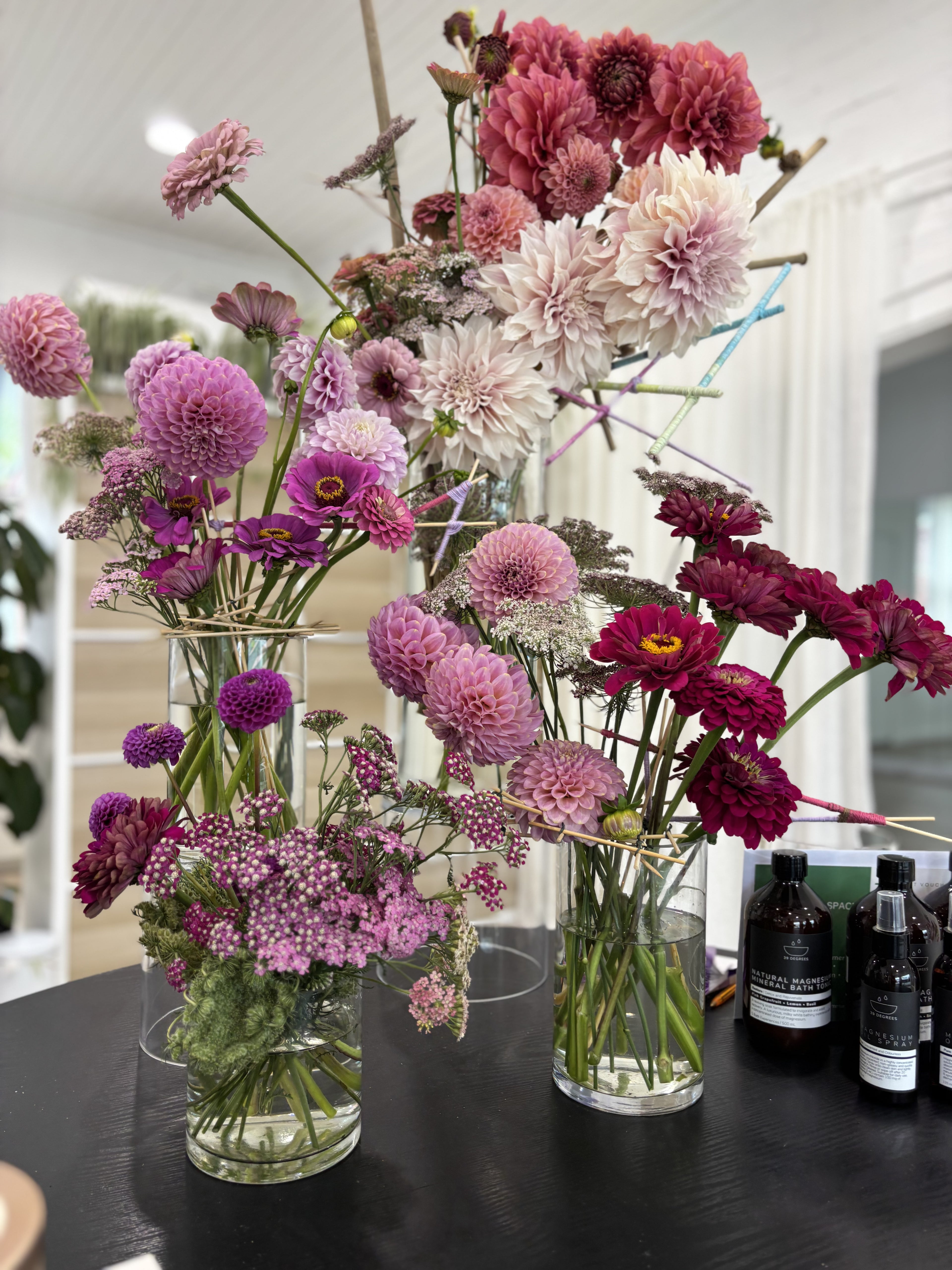 Bouquets of flowers in clear vases on a dark surface with bottles in the background.